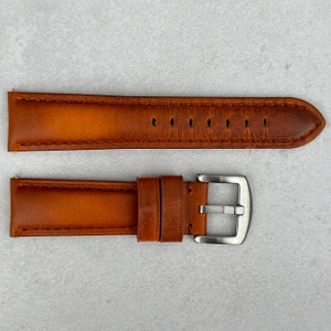 May include: A brown leather watch strap with a silver buckle. The strap has a stitched edge and a textured surface.