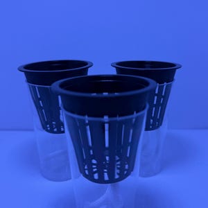 May include: Three black plastic net pots, designed for hydroponic gardening, are displayed. The pots have a cylindrical shape with slotted sides and sit inside clear plastic containers. The image is taken against a blue background.