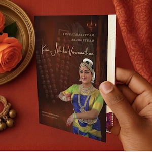 Può includere: Un opuscolo per un evento Bharatanatyam Arangetram, tenutosi il 13 novembre 2021, è tenuto in mano. La copertina dell'opuscolo mostra una donna in abito tradizionale della danza indiana. Rose arancioni e un piatto dorato a sinistra.