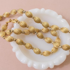 May include: A gold-toned necklace with ridged, oval beads. The necklace is resting on a white, scalloped-edge dish against a soft pink background. The clasp is visible, and the beads are strung together.