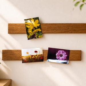 Könnte beinhalten: Zwei horizontale, rechteckige Holzleisten, die an einer Wand montiert sind und als magnetische Präsentationsschienen dienen. Die obere Leiste hält ein Foto eines gelben Ahornblatts, während die untere Leiste zwei Fotos, ein Feld und eine violette Blume, hält.
