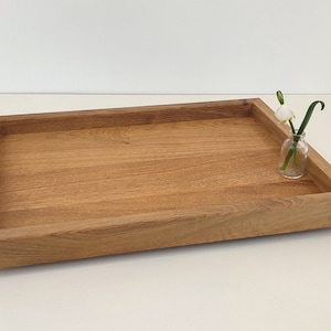 Könnte beinhalten: Ein rechteckiges Holztablett mit erhöhtem Rand aus hellbraunem Holz. Auf dem Tablett steht eine kleine Glasvase mit einer weißen Blume und einem grünen Stiel. Das Tablett ist leicht angewinkelt, wodurch seine klaren Linien und die natürliche Holzmaserung zur Geltung kommen.
