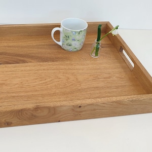 Może przedstawiać: Prostokątna drewniana taca do serwowania z dwoma uchwytami po bokach. Taca wykonana jest z jasnobrązowego drewna z naturalnym wykończeniem. Na tacy znajduje się biały kubek z kwiatowym wzorem i mały szklany wazon z pojedynczym białym kwiatem.