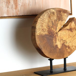 Op de afbeelding: Een houten sculptuur van een boomstamschijf met een natuurlijke houtkleur. Het beeld is gemonteerd op een zwarte metalen standaard met drie poten.