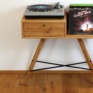 Op de afbeelding: Een houten draaitafelstandaard met een lade en een plank voor vinylplaten. De standaard heeft zwarte metalen poten en een natuurlijke houten afwerking.