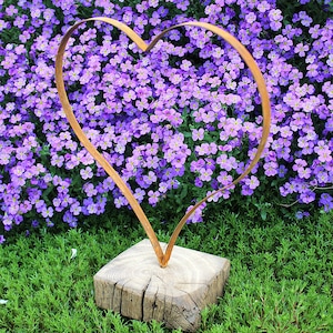 Op de afbeelding: Een bronzen metalen hartsculptuur op een vierkante houten basis. De sculptuur staat tegen een achtergrond van levendige paarse bloemen en groen gras. Het hartvormige ontwerp is eenvoudig en elegant.