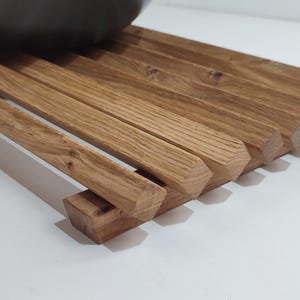 May include: A wooden trivet made of light brown wood with a natural finish. The trivet is made of multiple slats of wood that are arranged in a staggered pattern. The trivet is designed to protect surfaces from heat.