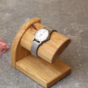 Pode incluir: Um suporte de relógio de madeira com um design curvo. O suporte é feito de madeira marrom claro e tem um acabamento liso. Um relógio prateado com um mostrador branco é exibido no suporte.