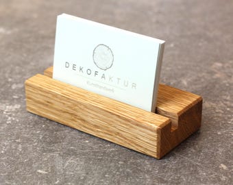 Könnte beinhalten: Ein Holz-Visitenkartenhalter mit natürlicher Holzoptik. Der Halter hat einen Schlitz für Visitenkarten und einen Sockel, um ihn aufrecht zu halten. Eine Visitenkarte mit dem Text "DEKOFAKTUR" und "Kunst handwerk" ist im Halter ausgestellt.