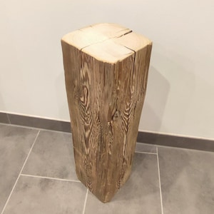 Könnte beinhalten: Ein hoher, quadratischer Holzsockel mit natürlicher, verwitterter Optik. Die Holzmaserung ist sichtbar und zeigt die Jahresringe des Baumes. Die Oberfläche ist hell, während die Seiten dunkler braun sind. Kann als Pflanzenständer oder Beistelltisch dienen.