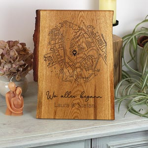 May include: A wooden plaque with a detailed map engraving. The plaque features a heart-shaped marker and text that reads "Wo alles begann Laura & Stefan". The wood has a natural grain and a rustic edge.