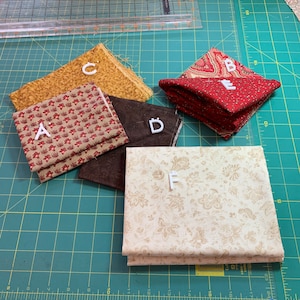 May include: A collection of six fabric squares with different patterns. The fabrics are in various colors, including brown, red, yellow, and white. The fabrics are arranged on a green cutting mat with a ruler and a white letter label for each fabric.