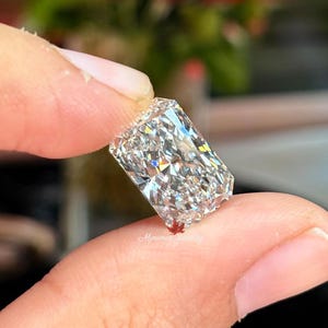 May include: A radiant-cut diamond held between fingers, showcasing its brilliant facets. The rectangular gemstone sparkles with internal reflections, reflecting light. The background is blurred, with hints of red and green.
