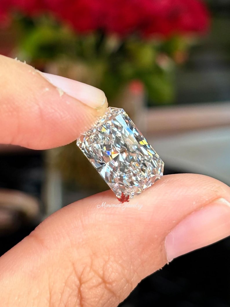 May include: A radiant-cut diamond held between fingers, showcasing its brilliant facets. The rectangular gemstone sparkles with internal reflections, reflecting light. The background is blurred, with hints of red and green.