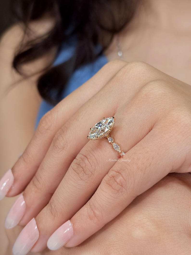 May include: A gold ring featuring a large marquise-cut center stone surrounded by a halo of smaller stones. The band is accented with additional marquise-cut stones along the sides. The text Minimal Jewelry is visible on the ring band.