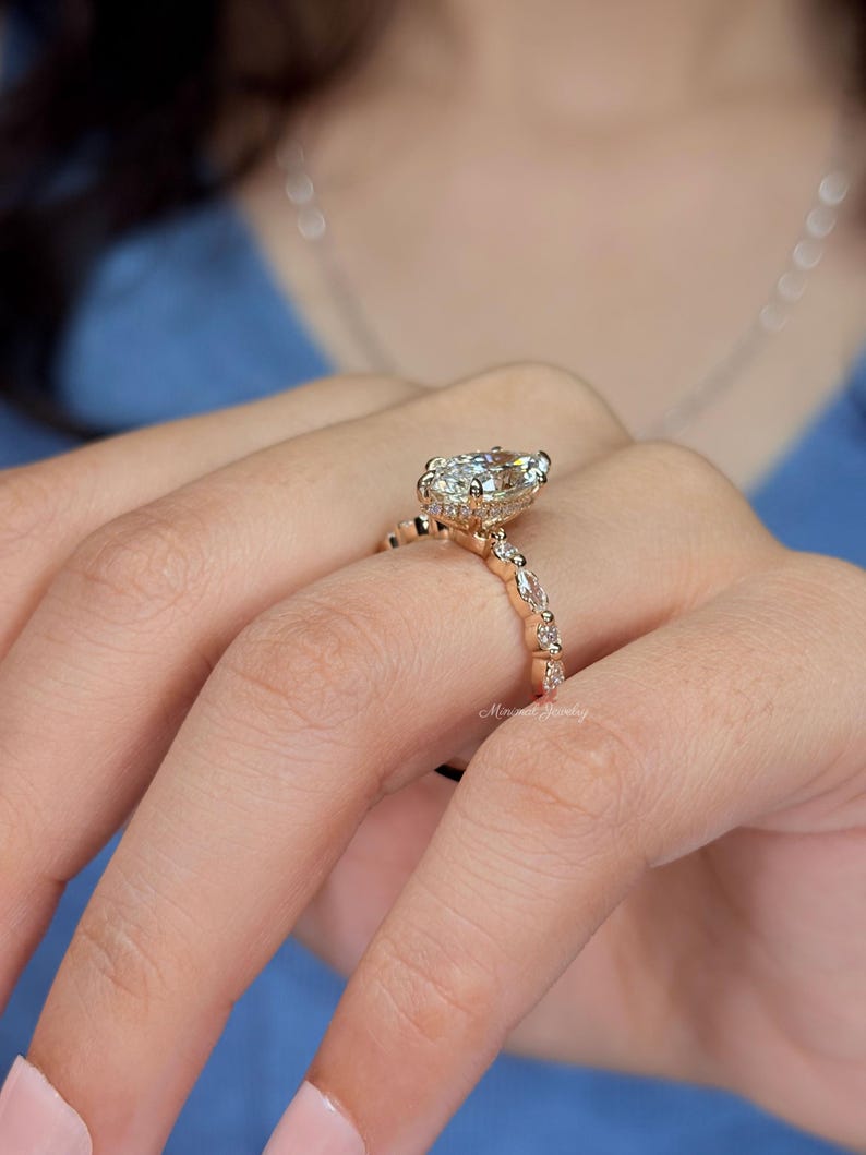 May include: A rose gold ring featuring a large pear-cut center stone and a band accented with alternating round and marquise-cut stones. The setting includes a hidden halo of small pav&eacute; stones beneath the main gem, with the text Minimal Jewelry.