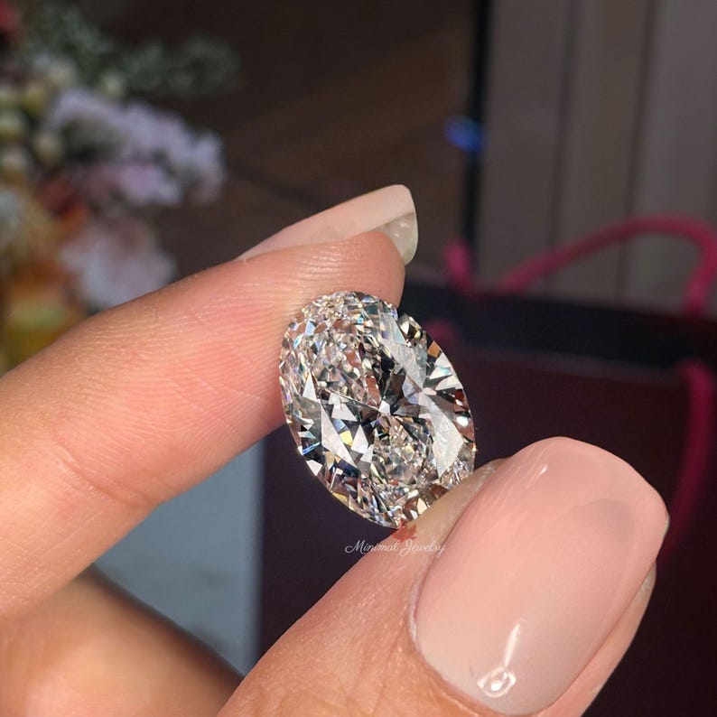 May include: A large, oval-cut diamond with brilliant facets, held between fingers. The diamond sparkles with internal reflections, showcasing its clarity and cut. The background is blurred, focusing attention on the gemstone. The image is well-lit, highlighting the diamond's brilliance.