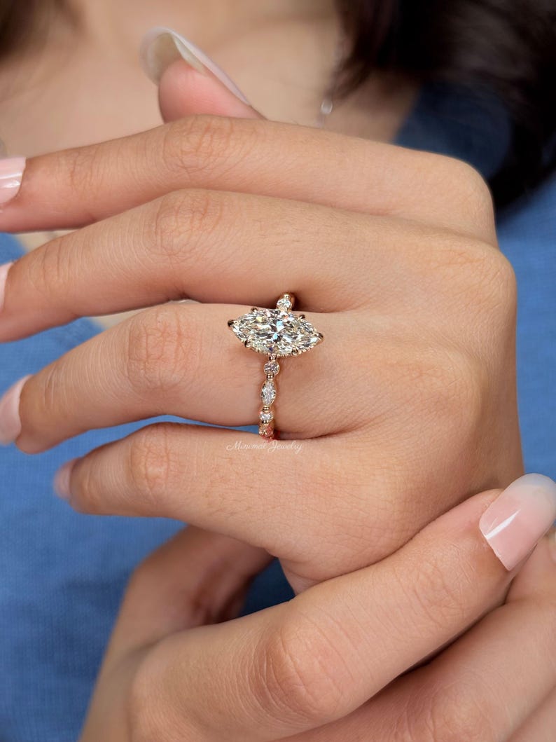 May include: A rose gold ring featuring a large marquise-cut center stone held by prongs. The band is adorned with a series of smaller round and marquise-cut stones set in a delicate, repeating pattern along the shank, creating a refined appearance.
