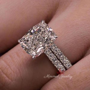 Könnte beinhalten: Ein silberfarbener Ring mit einem großen, rechteckigen, facettierten Mittelstein, der von kleineren, funkelnden Steinen umgeben ist. Die Ringschiene ist zudem mit einer Reihe kleiner, klarer Brillantschliff-Steine besetzt. Der Text Minimal Jewelry ist sichtbar.