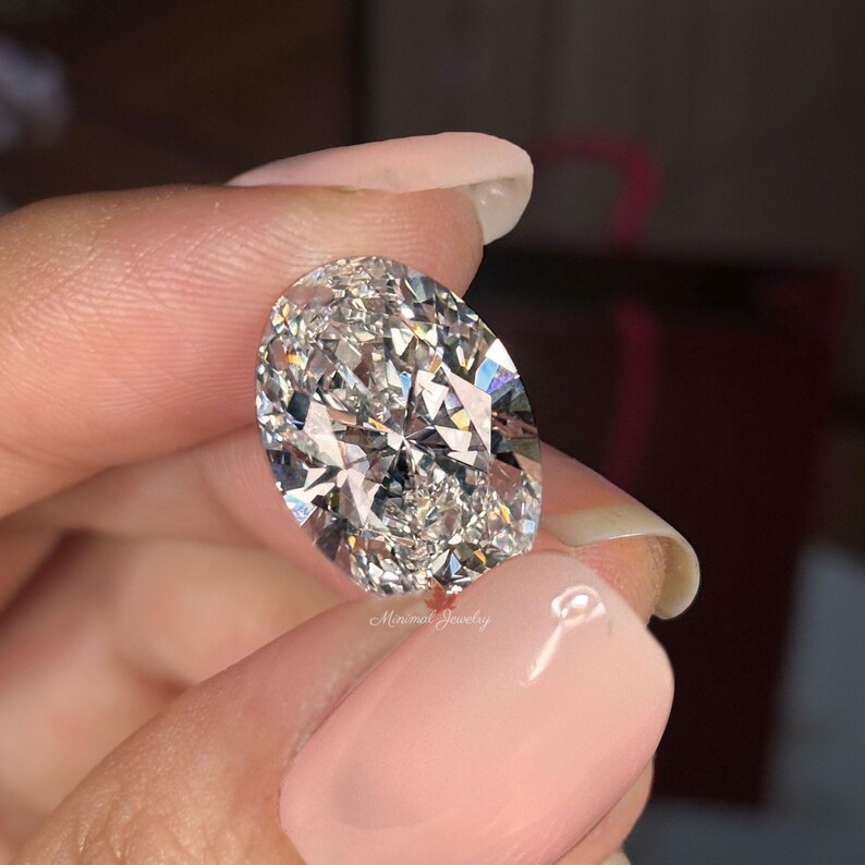 May include: A large, oval-cut diamond with brilliant facets, held between fingers. The diamond sparkles with internal reflections, showcasing its clarity and cut. The background is blurred, focusing attention on the gemstone. The image is well-lit, highlighting the diamond's brilliance.