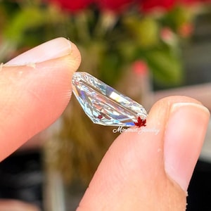 May include: A clear, elongated gemstone with multiple facets, held between fingers. The gem reflects light, displaying a spectrum of colors. The background features blurred red flowers and greenery. The text "Minimal Jewelry" is visible on the gem.