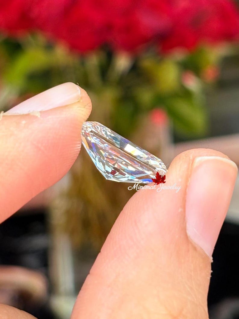May include: A clear, elongated gemstone with multiple facets, held between fingers. The gem reflects light, displaying a spectrum of colors. The background features blurred red flowers and greenery. The text "Minimal Jewelry" is visible on the gem.