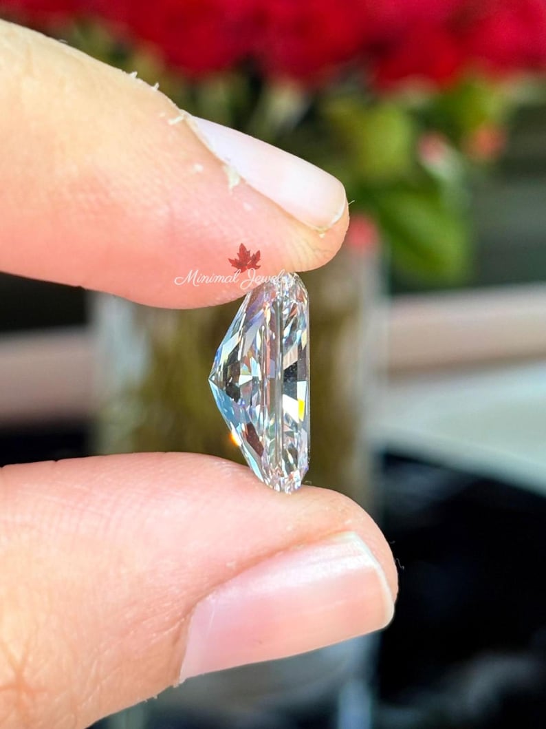 May include: A clear, rectangular-cut gemstone held between fingers. The gem has multiple facets that reflect light, creating a sparkling effect. The image is close-up, highlighting the clarity and cut of the stone. The text "Minimal Jewels" is visible.