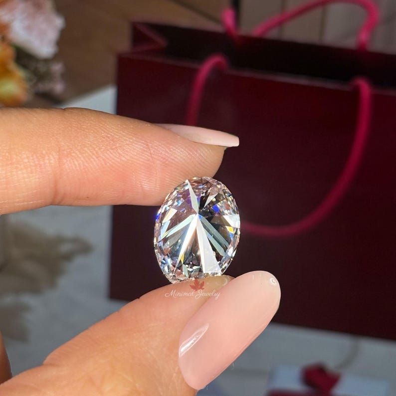 May include: A brilliant, oval-cut diamond with multiple facets, held between fingers. The gemstone sparkles with light, showcasing its clarity and cut. A red gift bag is in the background. The image is well-lit, highlighting the diamond's brilliance.