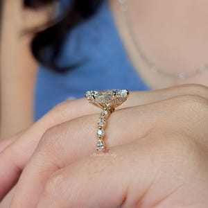 May include: A gold ring featuring a large, elongated marquise-cut center stone set horizontally. The band is adorned with a series of smaller, alternating round and pear-shaped diamonds, and a hidden halo of tiny stones encircles the base of the setting.