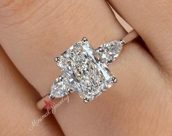 May include: A three-stone ring featuring a large rectangular radiant-cut center diamond flanked by two smaller pear-cut side diamonds on a thin polished metal band. The ring includes a small red leaf logo and the text Minimal Jewelry.