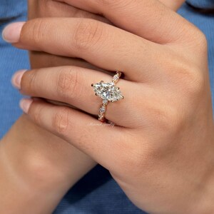May include: A gold ring featuring a large marquise-cut center stone and smaller round-cut stones along the band. The metal band has a warm gold tone, and the stones are clear and faceted. The text Minimal Jewelry is visible on the ring band.