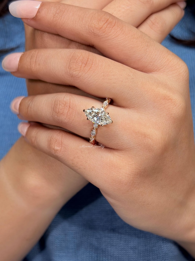 May include: A gold ring featuring a large marquise-cut center stone and smaller round-cut stones along the band. The metal band has a warm gold tone, and the stones are clear and faceted. The text Minimal Jewelry is visible on the ring band.