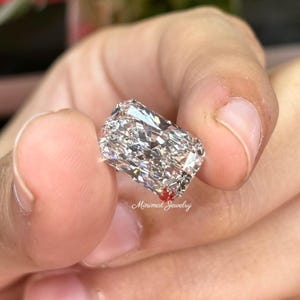 May include: A large, rectangular-cut diamond with brilliant facets, held between fingers. The diamond sparkles with internal reflections, showcasing its clarity and cut. The image includes the text "Minimal Jewelry".
