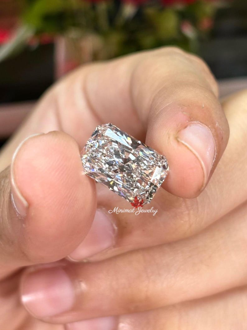 May include: A large, rectangular-cut diamond with brilliant facets, held between fingers. The diamond sparkles with internal reflections, showcasing its clarity and cut. The image includes the text "Minimal Jewelry".