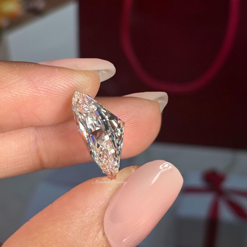 May include: A clear, faceted diamond with a unique, asymmetrical shape. The diamond is held between fingers, showcasing its brilliance and clarity. The background is a blurred red.