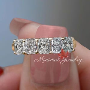 Könnte beinhalten: Ein goldener Ring mit fünf Diamanten im Radiant-Schliff, eng nebeneinander gefasst. Die Diamanten funkeln hell. Der Ring wird vor einem unscharfen Hintergrund gehalten. Der Text "Minimal Jewelry" ist sichtbar.