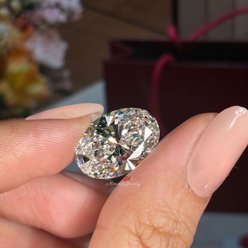 May include: A large, oval-cut diamond with brilliant facets, held between fingers with a light pink nail polish. The diamond sparkles with internal reflections. The background includes a blurred floral arrangement and a red box.