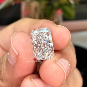 May include: A rectangular-cut diamond with a brilliant, multifaceted surface. The diamond is held between fingers, showcasing its clarity and sparkle. The image is well-lit, highlighting the gem's brilliance. The text "Minimal Jewelry" is visible.