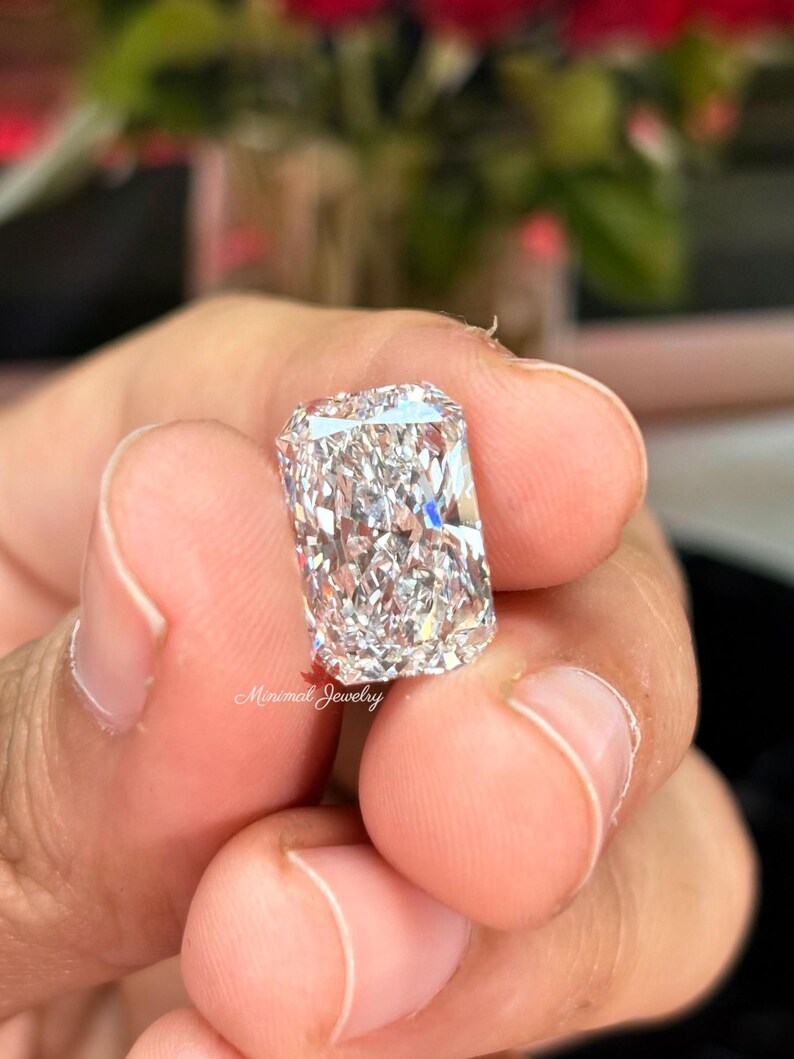 May include: A rectangular-cut diamond with a brilliant, multifaceted surface. The diamond is held between fingers, showcasing its clarity and sparkle. The image is well-lit, highlighting the gem's brilliance. The text "Minimal Jewelry" is visible.