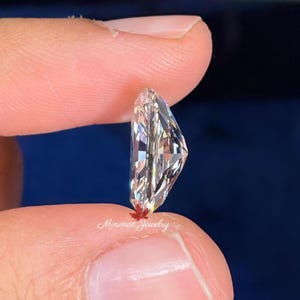 May include: A clear, faceted gemstone held between two fingers. The gemstone has a unique, asymmetrical shape with multiple facets that reflect light. The text "Minimal Jewelry" is visible near the base of the stone.
