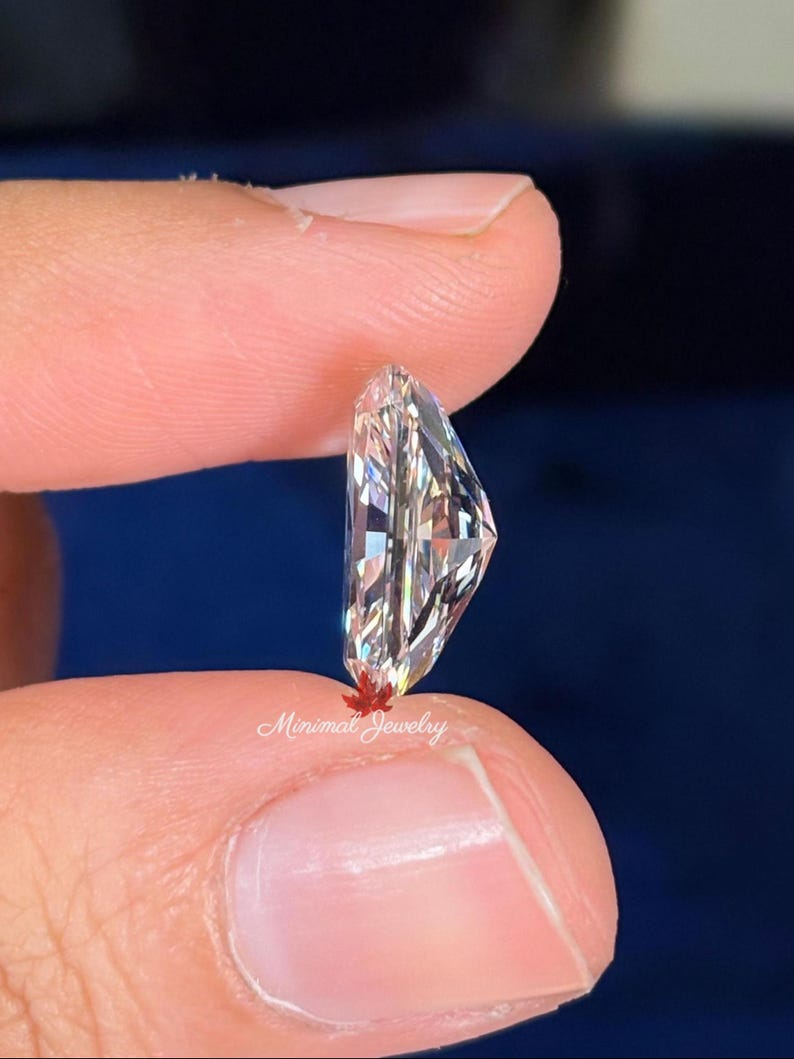May include: A clear, faceted gemstone held between two fingers. The gemstone has a unique, asymmetrical shape with multiple facets that reflect light. The text "Minimal Jewelry" is visible near the base of the stone.