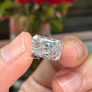 May include: A rectangular-cut diamond held between fingers. The diamond is clear and multifaceted, reflecting light. The background features blurred red flowers and the text "Minimal Jewelry".