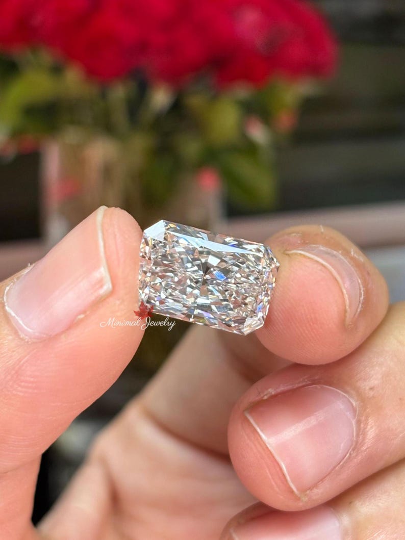 May include: A rectangular-cut diamond held between fingers. The diamond is clear and multifaceted, reflecting light. The background features blurred red flowers and the text "Minimal Jewelry".