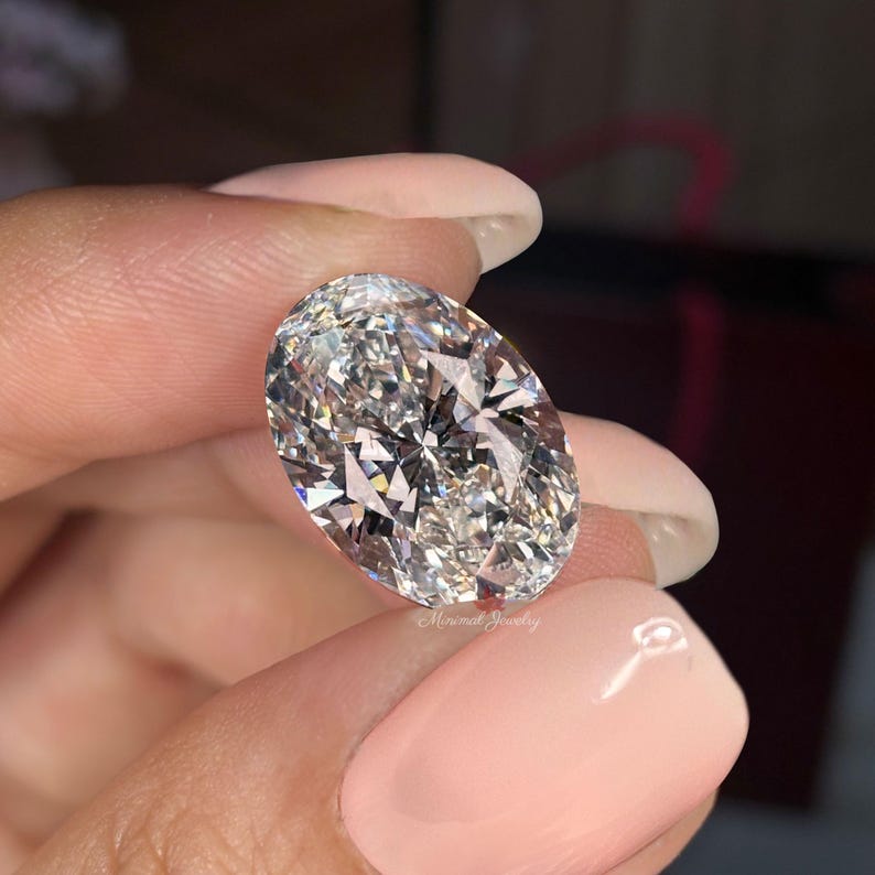 May include: A large, oval-cut diamond held between fingers. The diamond is clear and reflects light, showcasing numerous facets. The image is a close-up, highlighting the stone's brilliance and clarity. The text "Minimal Jewelry" is visible.