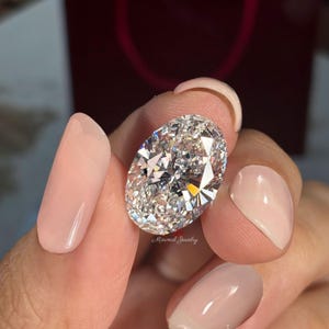 May include: A large, oval-cut diamond with brilliant facets, held between fingers. The diamond sparkles with light reflections. The text "Minimal Jewelry" is visible. The nails are painted with a light pink polish.