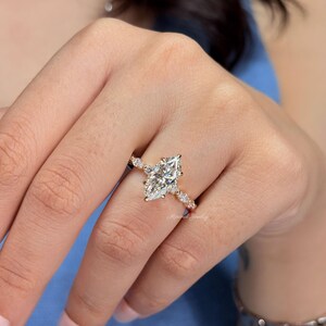 May include: A gold ring featuring a large marquise-cut center stone and smaller round-cut stones along the band. The ring has a delicate metal setting and is worn on a finger. The text Minimal Jewelry is visible on the band of the ring.