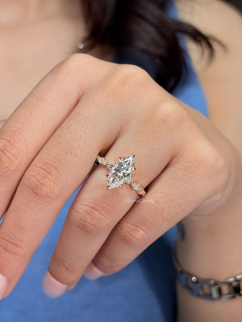 May include: A gold ring featuring a large marquise-cut center stone and smaller round-cut stones along the band. The ring has a delicate metal setting and is worn on a finger. The text Minimal Jewelry is visible on the band of the ring.