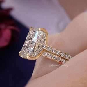 May include: A gold ring featuring a large rectangular radiant-cut center stone held by four prongs. The band and the basket setting are encrusted with small round pavé diamonds. The text Minimal Jewelry is visible on the lower right side.