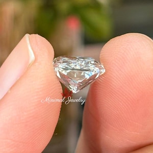 May include: A clear, faceted diamond held between two fingers. The diamond is cut with multiple facets, reflecting light. The text "Minimal Jewelry" is visible in the image. The background is blurred, with hints of green and red.