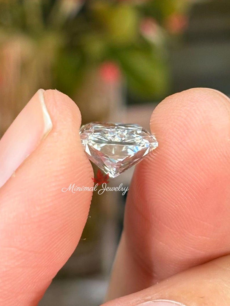 May include: A clear, faceted diamond held between two fingers. The diamond is cut with multiple facets, reflecting light. The text "Minimal Jewelry" is visible in the image. The background is blurred, with hints of green and red.